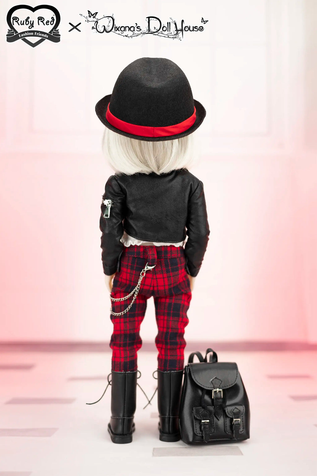 Doll wearing a black outfit with red plaid pants, black hat, and black boots, standing next to a black backpack on a pink background.