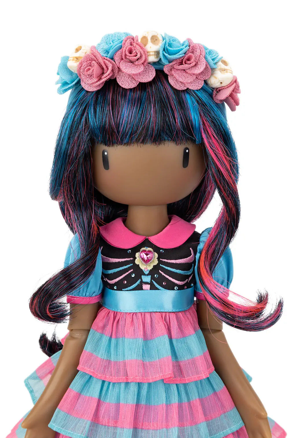 A stylized doll with long blue, pink, and black streaked hair, wearing a floral headband with small cream skulls and a pink-and-blue dress with a striped ruffled skirt, shown against a plain white background.