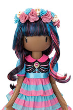 A stylized doll with long blue, pink, and black streaked hair, wearing a floral headband with small cream skulls and a pink-and-blue dress with a striped ruffled skirt, shown against a plain white background.