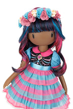 A stylized doll with dark brown skin, long wavy blue and pink hair, a floral headband with flowers and skull motifs, and a layered dress in pink and light blue with a black bodice, shown on a plain white background.