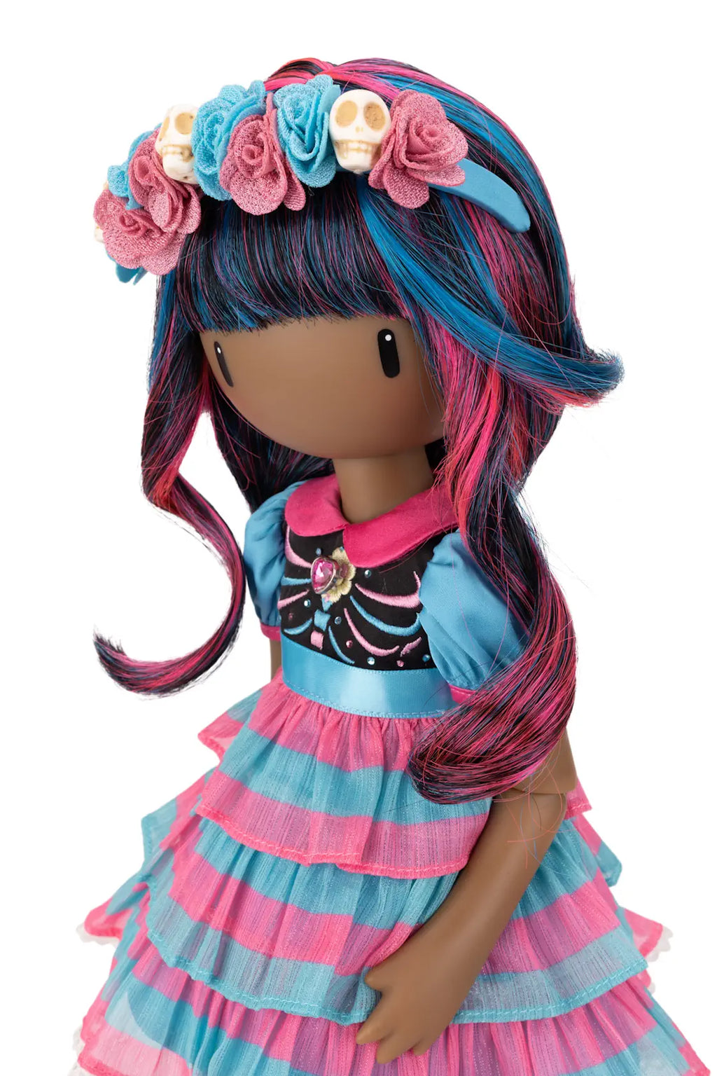 A doll with dark brown skin and long, wavy hair in black, blue, and pink, wearing a floral headband with pink and blue roses and skull details, a black top with a pink collar, and a tiered blue and pink dress, shown against a plain white background.
