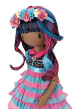 A doll with dark brown skin and long, wavy hair in black, blue, and pink, wearing a floral headband with pink and blue roses and skull details, a black top with a pink collar, and a tiered blue and pink dress, shown against a plain white background.