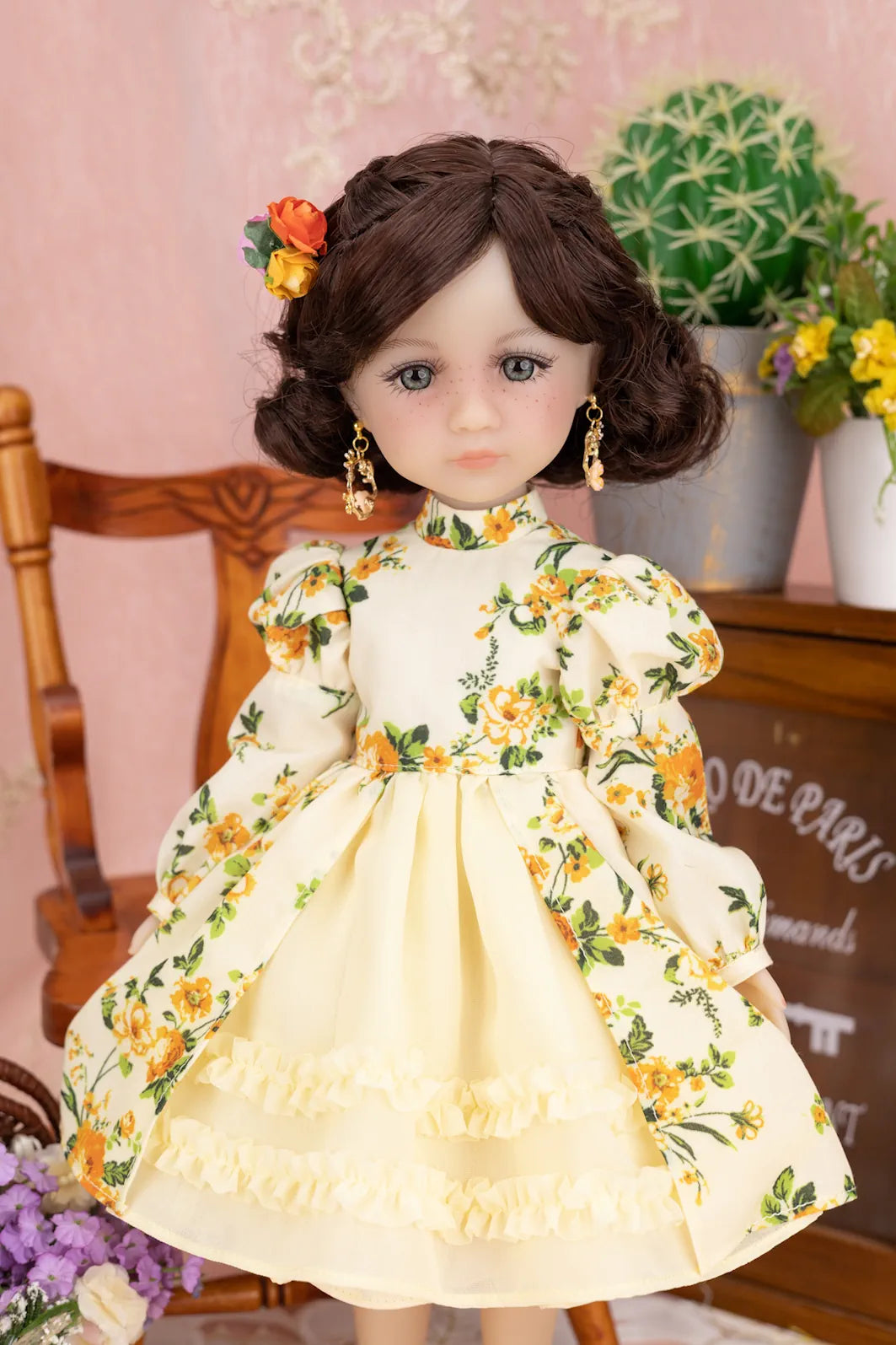 Doll in a yellow floral dress with a decorative headpiece against a pink wall.