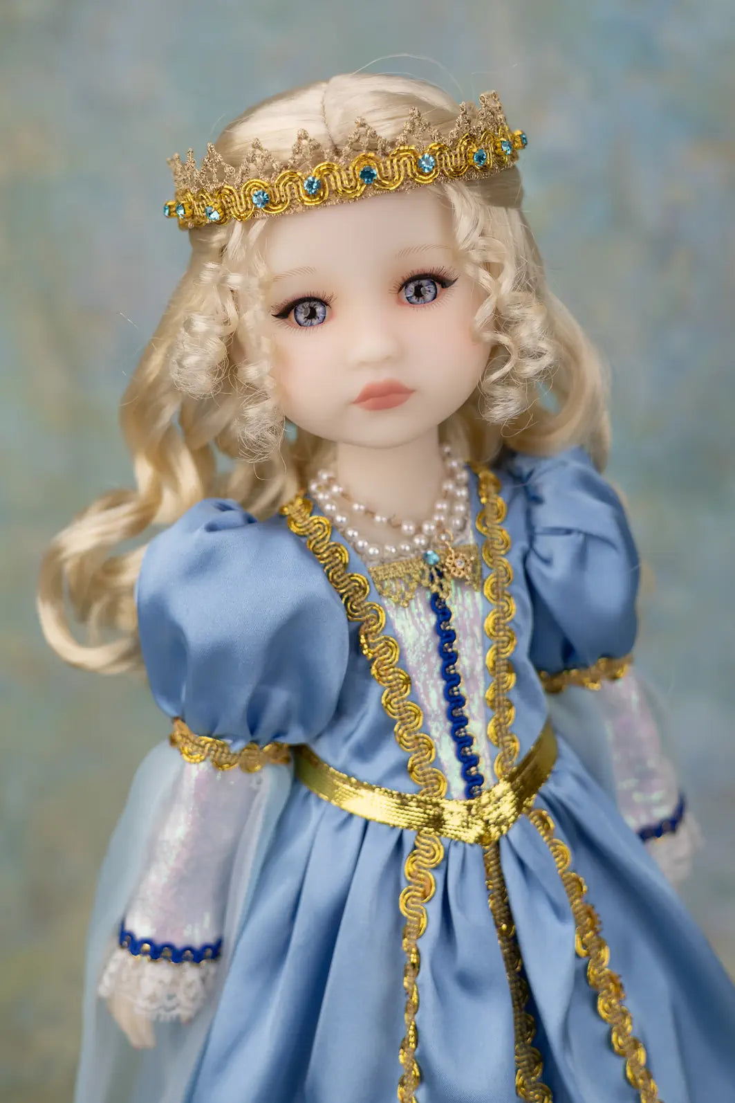 A close-up of a blonde doll wearing a light blue gown with gold trim, a pearl necklace, and a gold crown with blue stones, set against a soft blue background.