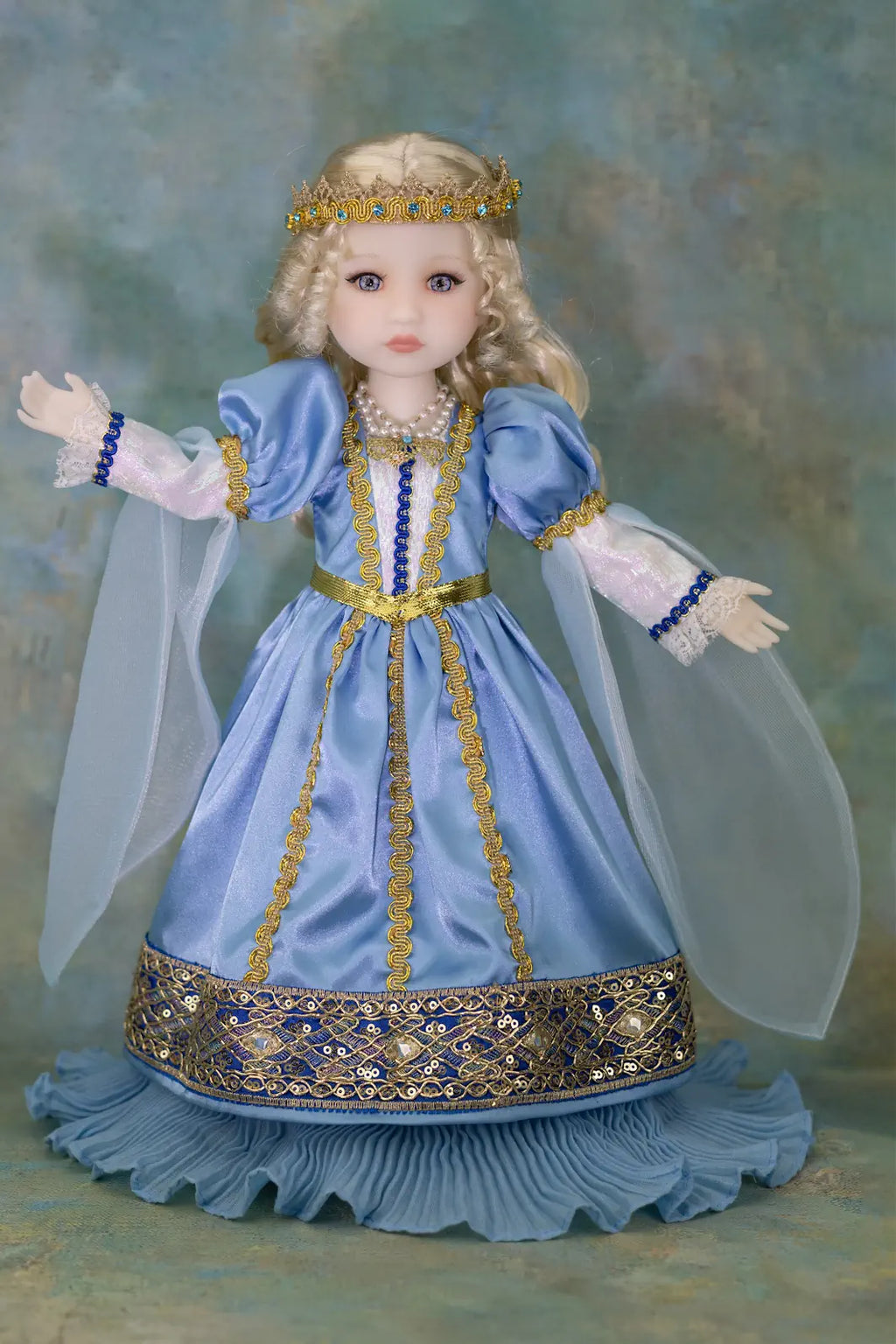 A doll dressed in a light blue medieval-style gown with gold trim, white sleeves, and a matching gold crown, posed against a textured blue-green backdrop.