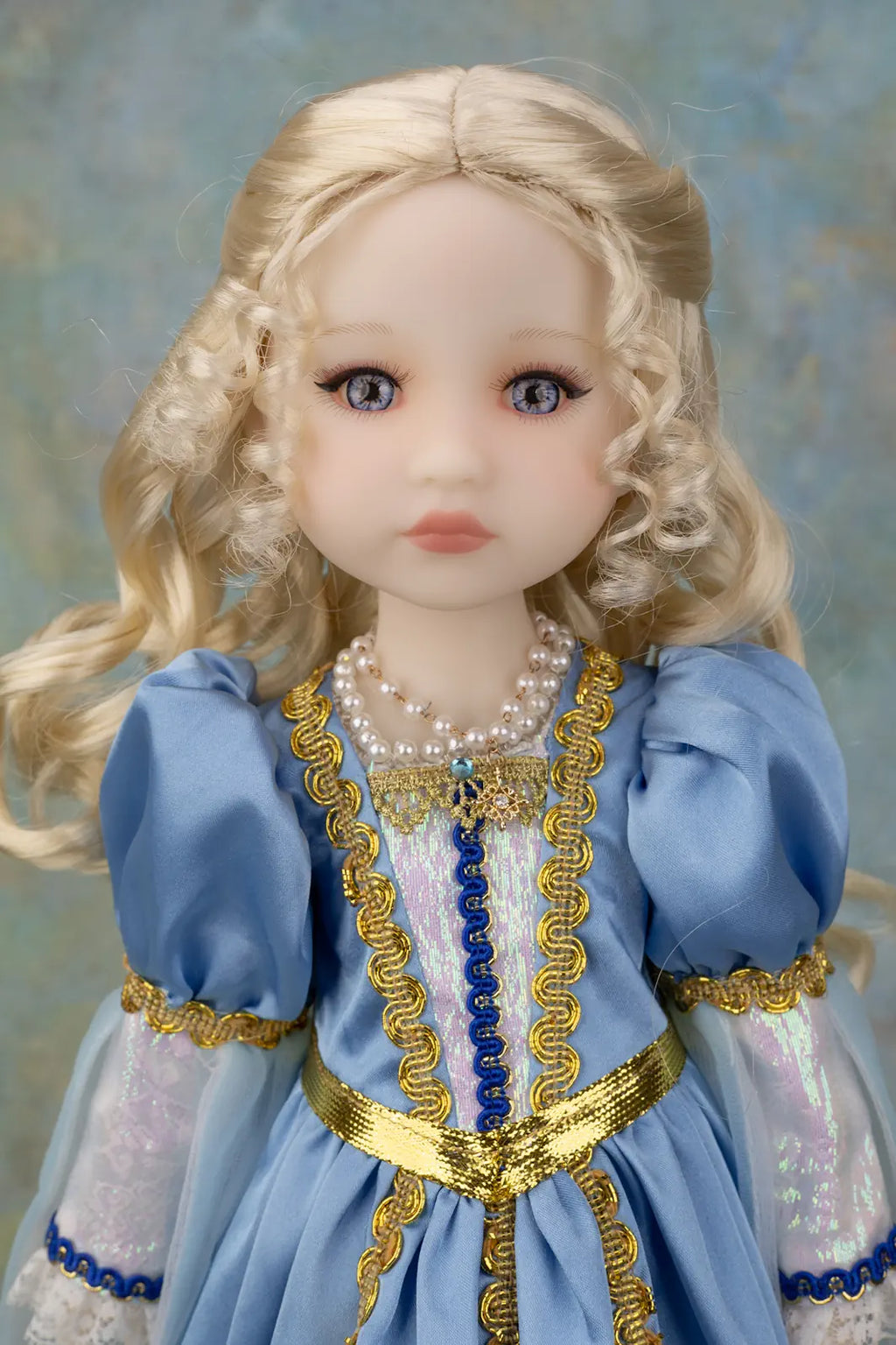 A close-up of a doll with long blonde curly hair, blue eyes, and a light blue dress with gold trim and pearl necklaces, set against a soft blue textured background.