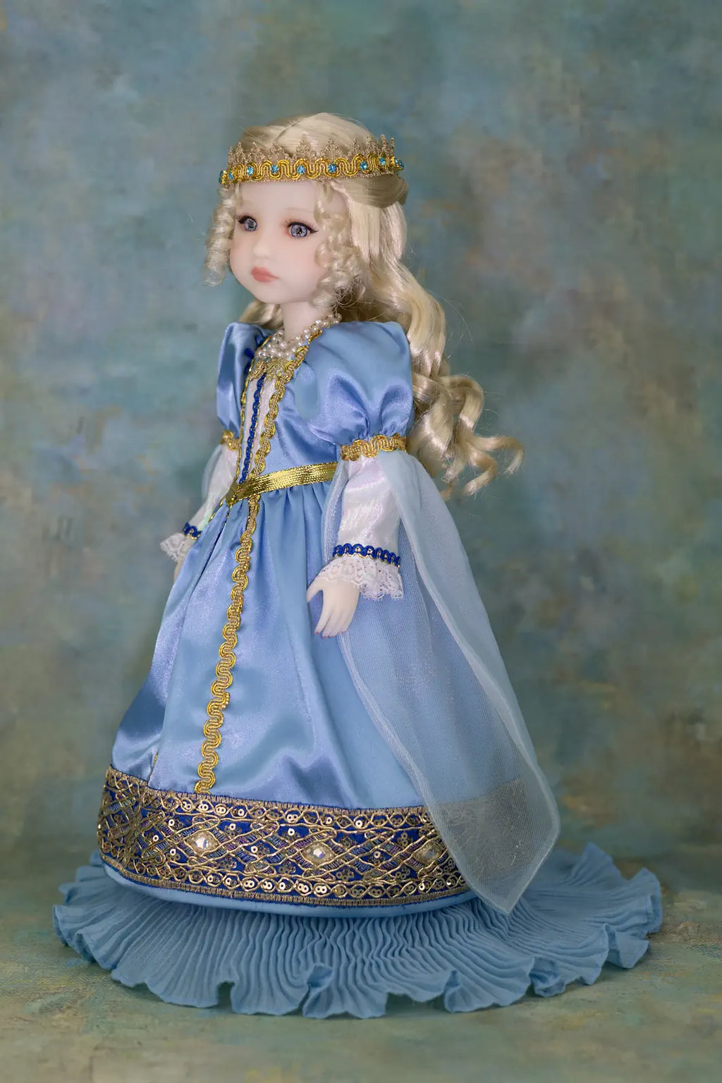 A blonde doll with a tiara wearing a light blue medieval-style gown with gold embroidery and a sheer cape, posed against a textured blue-green studio backdrop.