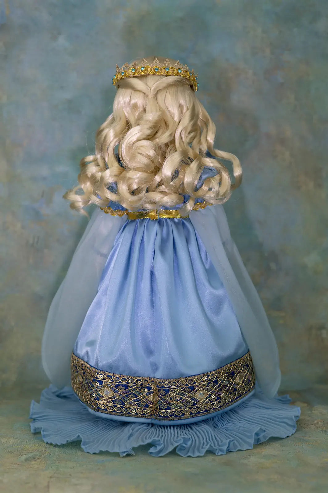 A doll  viewed from the back, wearing a long blonde wig, a gold crown, a light blue dress with gold trim, and a sheer blue veil, set against a textured blue studio background.