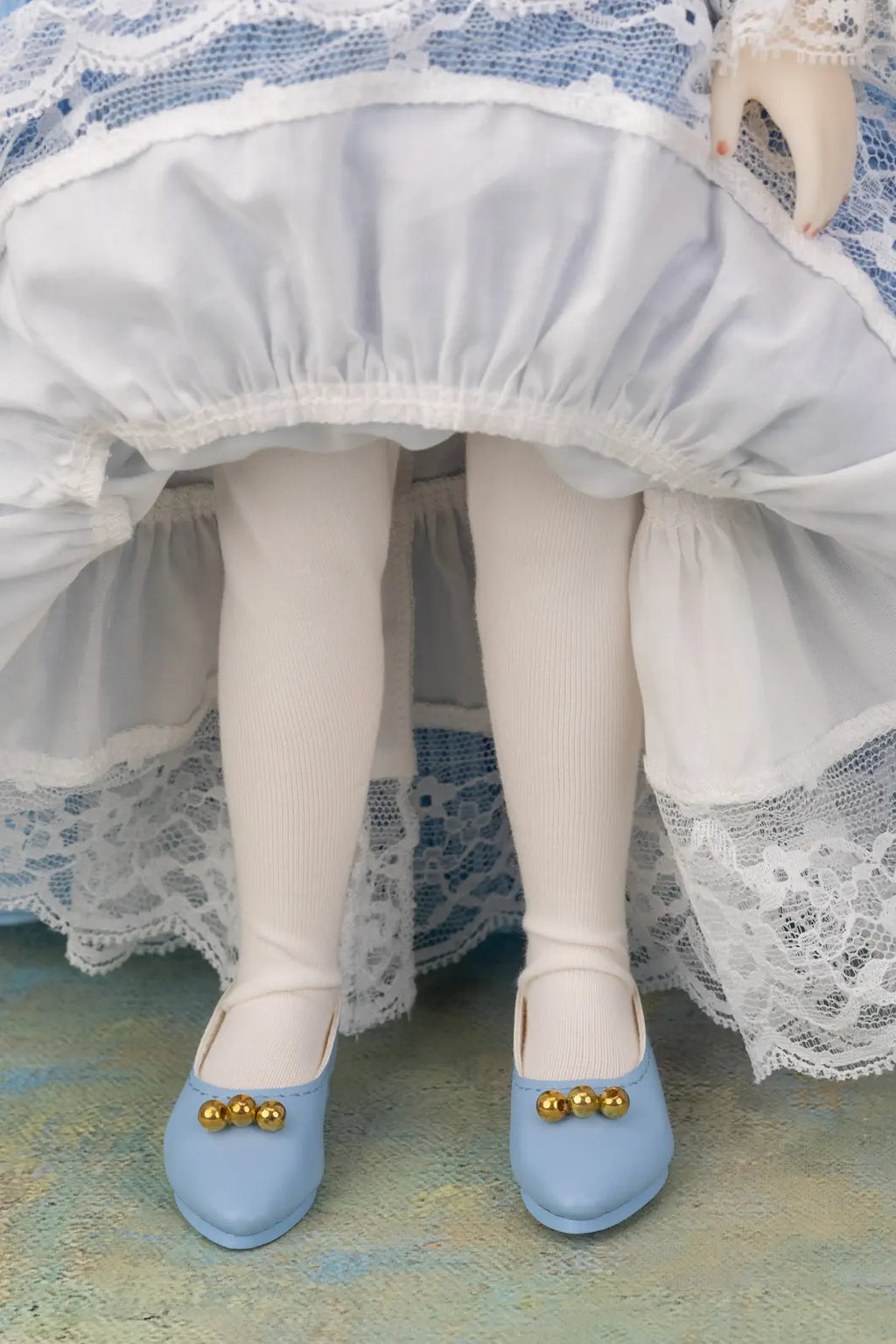 A close-up of a doll's legs wearing white tights and light blue shoes with gold decorative beads, set against a white and blue ruffled dress.