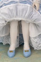 A close-up of a doll's legs wearing white tights and light blue shoes with gold decorative beads, set against a white and blue ruffled dress.