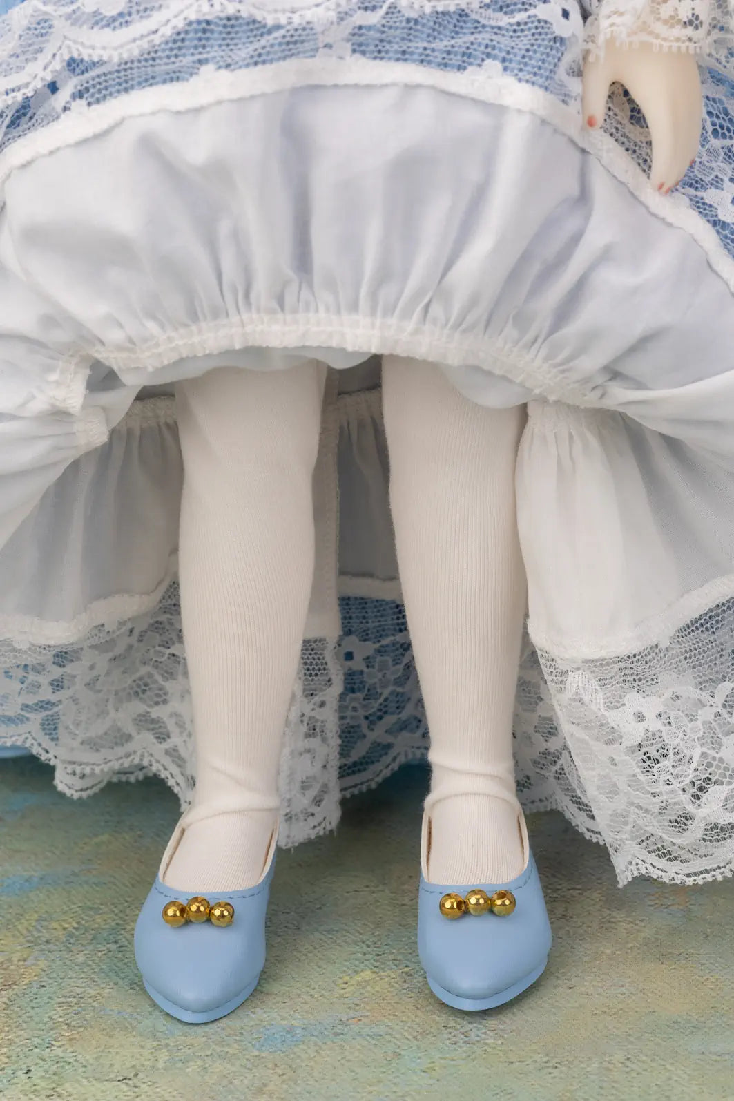 A close-up of a doll's legs wearing white tights and light blue shoes with gold decorative beads, set against a white and blue ruffled dress.