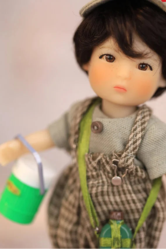 Boy doll wearing plaid suspender overalls with a tiny green lunch flask can in hand and a pale muted background