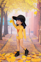 Doll in a yellow outfit with a black hat standing on a leaf-covered street with autumn trees.
