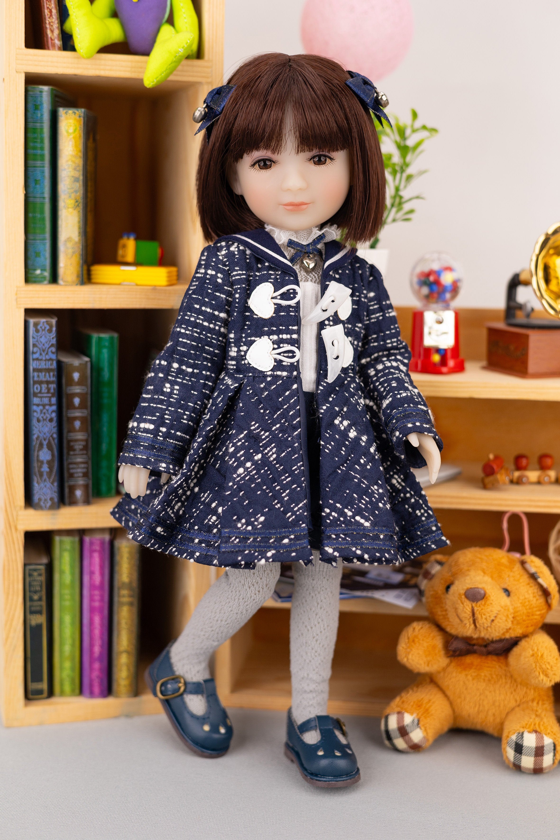 Doll in a blue coat standing next to a teddy bear in a room with books and toys.