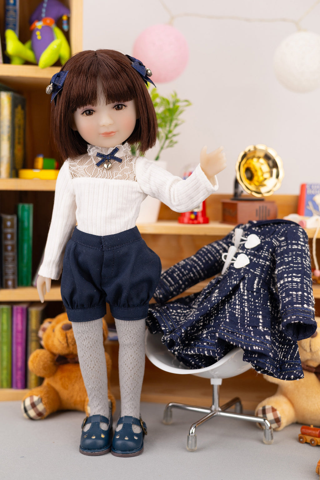Doll in a white blouse and navy pants standing next to a chair with a patterned dress on it, in a room setting.