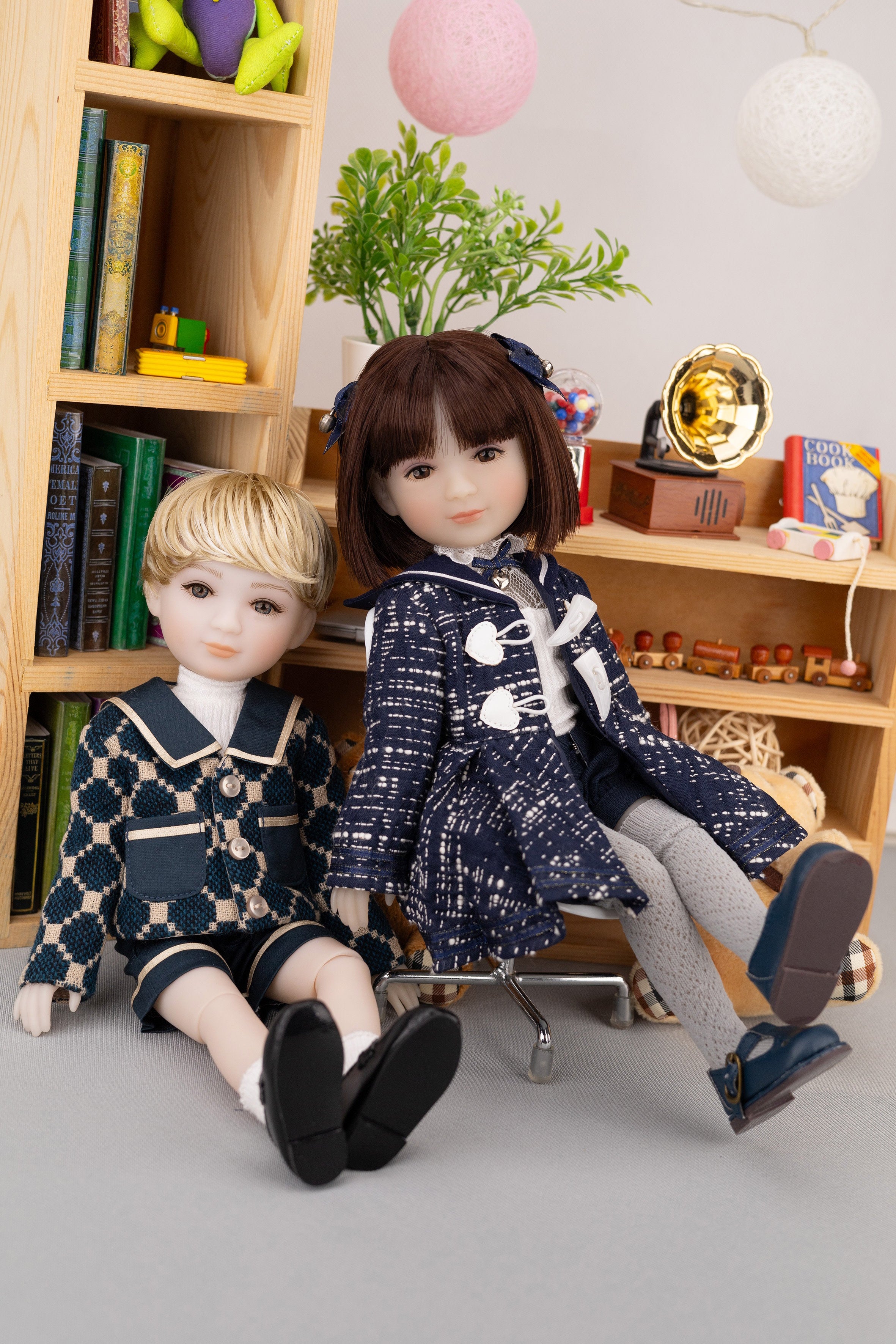 Two dolls in school uniforms sitting on a chair in front of a bookshelf.