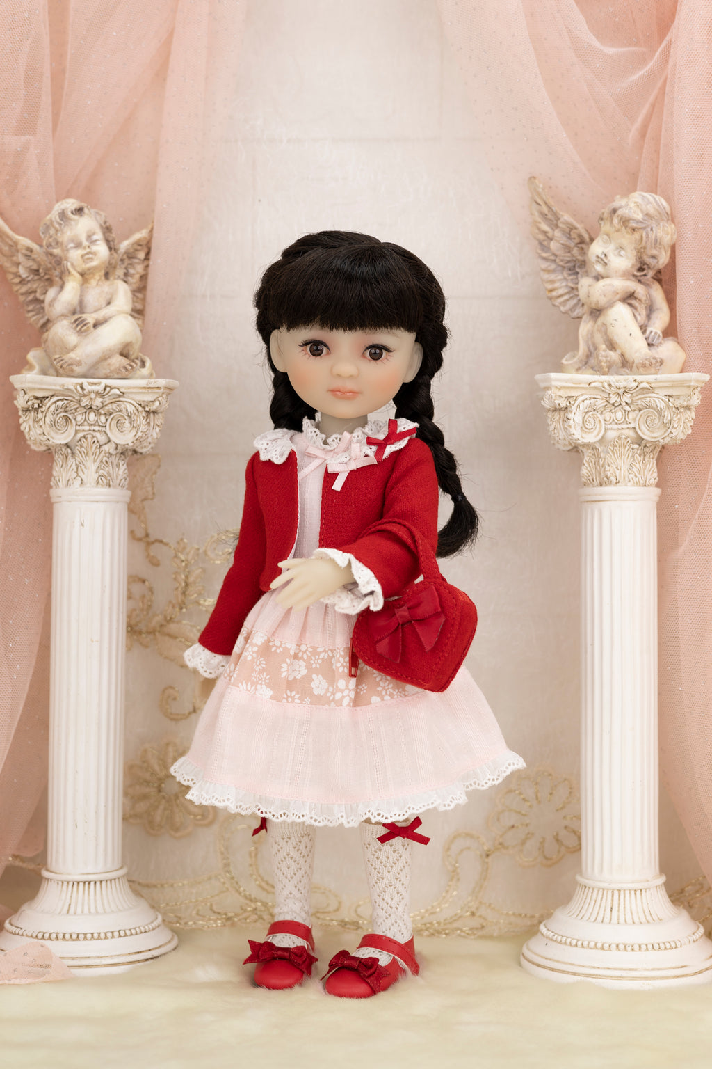 Doll in a red outfit standing between two decorative columns with angel statues.