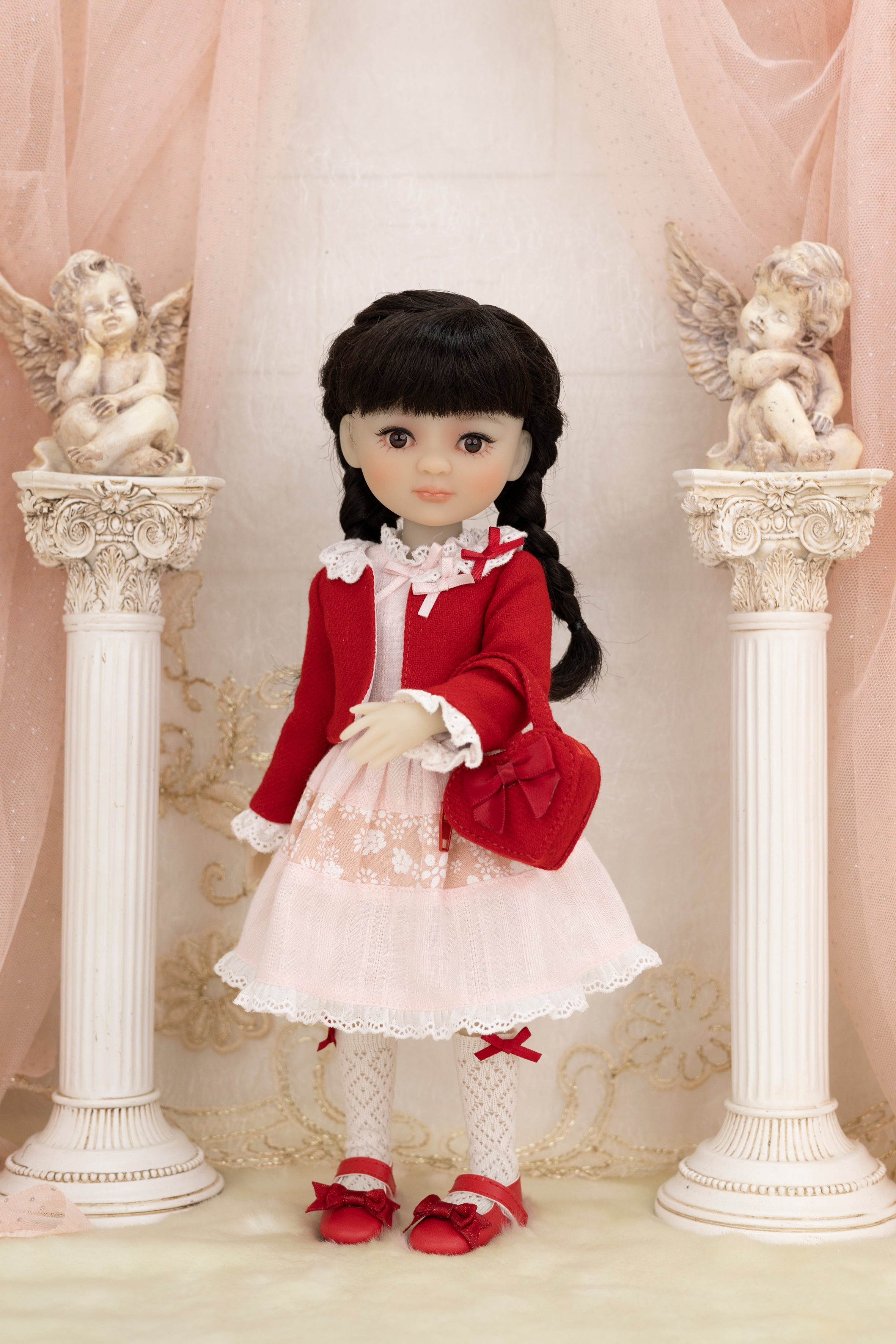 Doll in a red outfit standing between two decorative columns with angel statues.