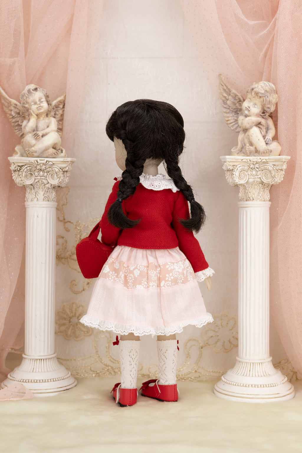 Doll in a red sweater and white dress standing between two decorative columns with angel statues.