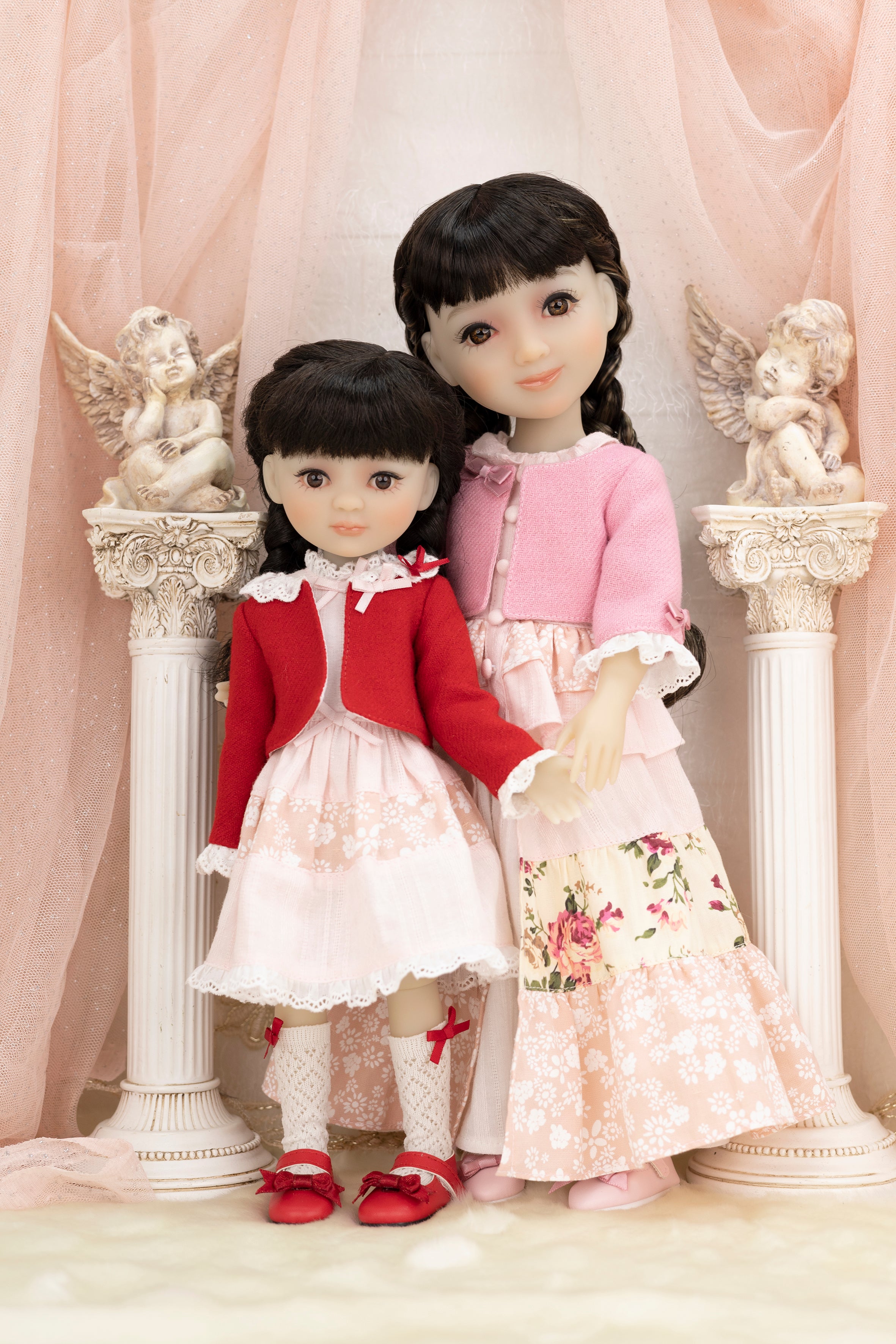 Two dolls in formal attire standing in front of decorative columns with angel statues.