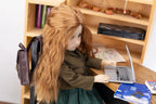 Doll with long hair using a laptop at a desk with books and a backpack.