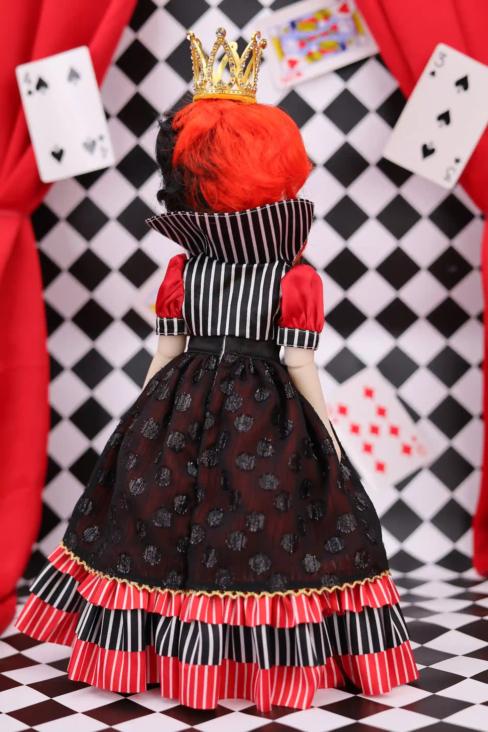 Doll in a red and black outfit with a crown, standing against a checkered and card-themed background.