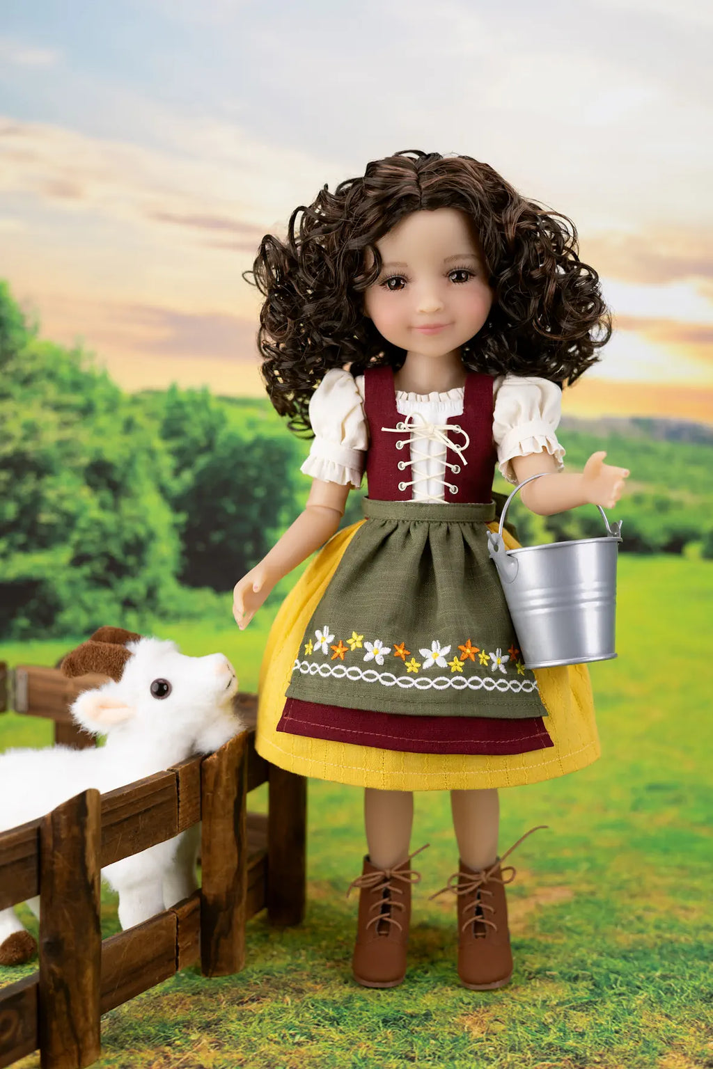 A doll with curly brown hair stands in a stylized outdoor farm scene, wearing a traditional dirndl dress with white sleeves, a burgundy bodice, olive green apron, yellow skirt, and brown lace-up boots. The doll holds a small silver bucket and stands beside a wooden fence with a white sheep toy.