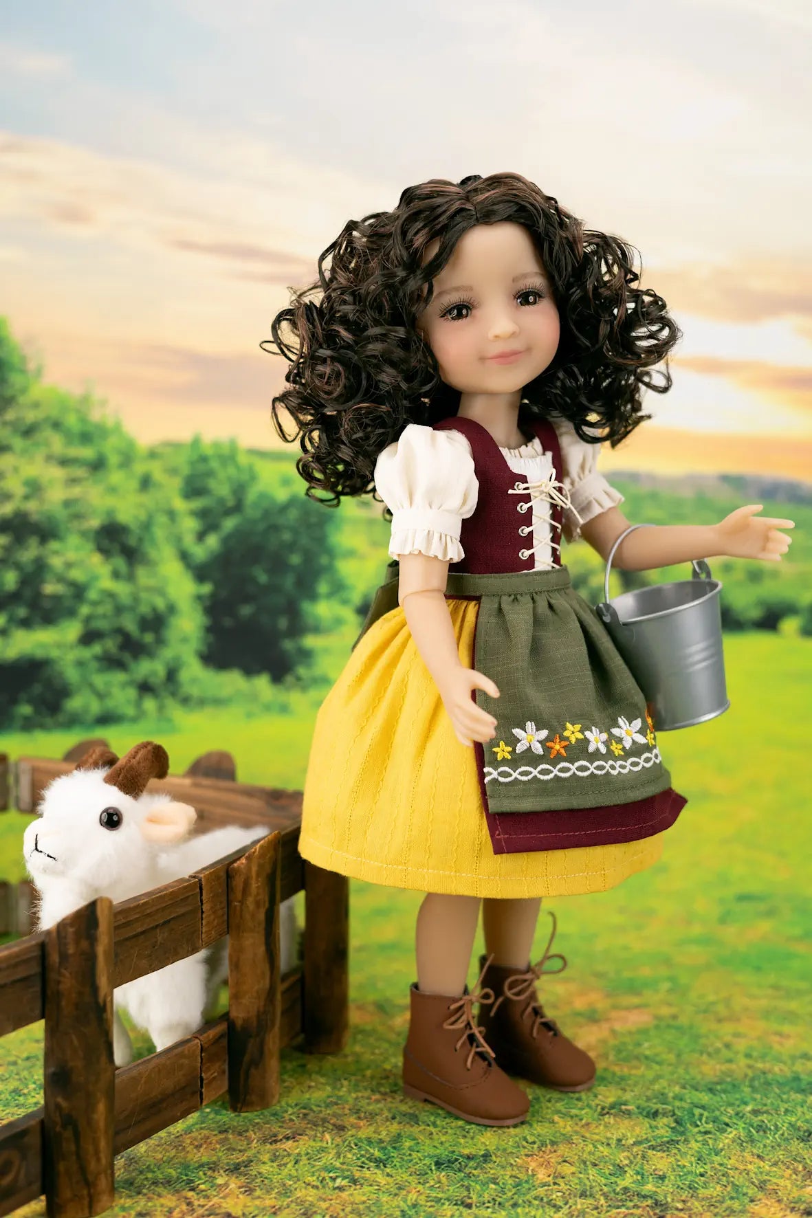 A doll with curly brown hair wearing a traditional German-style outfit with a yellow skirt, green apron, and white blouse, standing in a grassy field next to a small white sheep in a wooden pen.