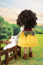 A doll viewed from behind wearing a voluminous dark brown curly wig, a yellow skirt, a white top, a green bow, and brown boots, standing in a grassy outdoor scene next to a wooden fence and a white plush sheep.
