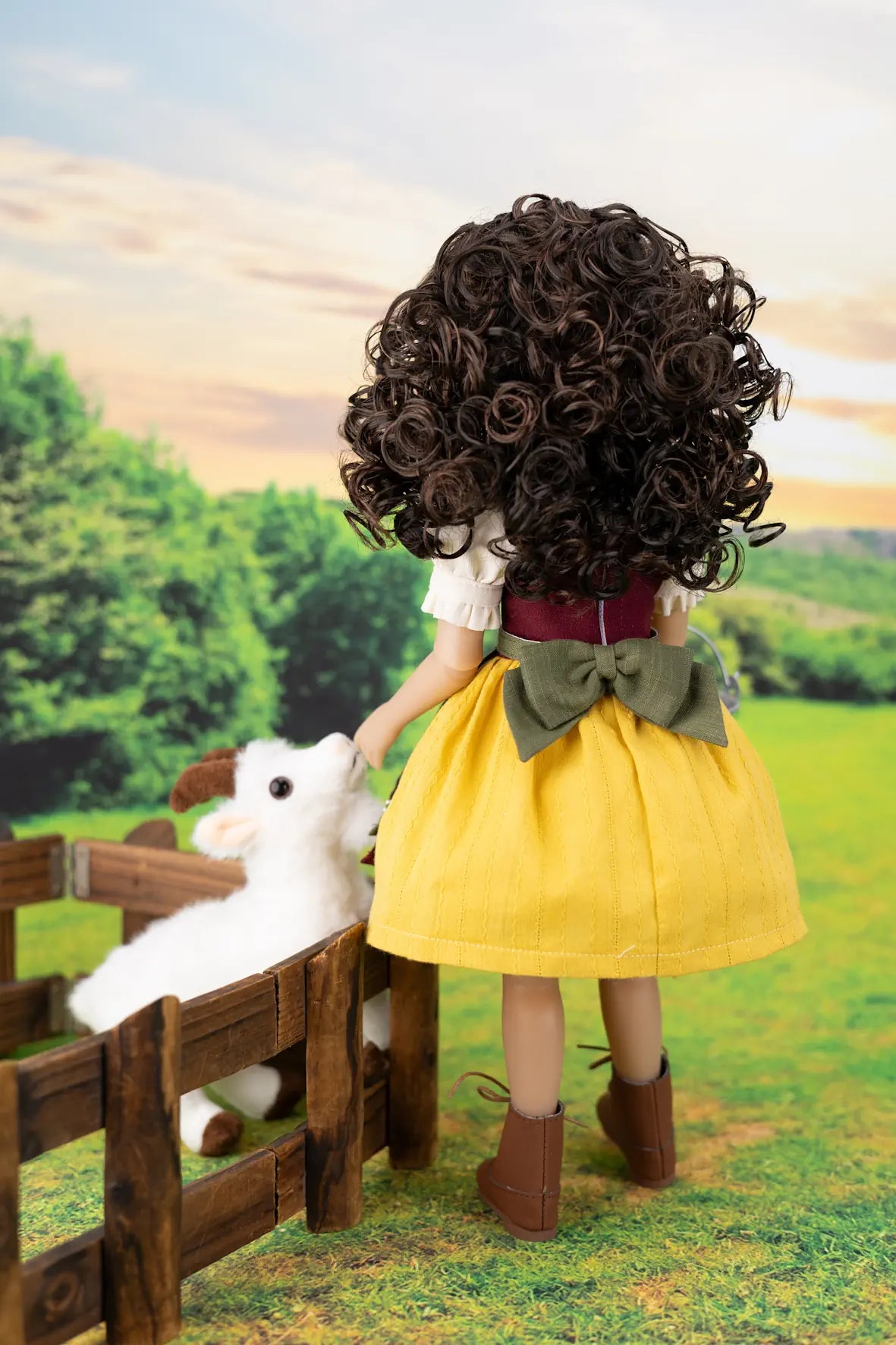 A doll viewed from behind wearing a voluminous dark brown curly wig, a yellow skirt, a white top, a green bow, and brown boots, standing in a grassy outdoor scene next to a wooden fence and a white plush sheep.