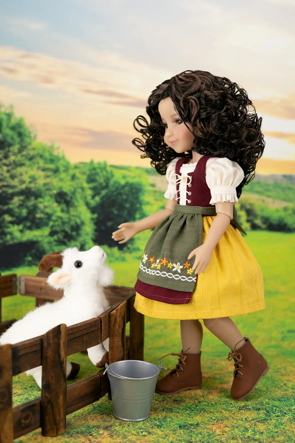 A doll with long curly brown hair stands beside a small wooden fence in a stylized outdoor farm scene, reaching toward a white plush lamb while wearing a traditional-style dress with a yellow skirt, green apron, and brown boots.