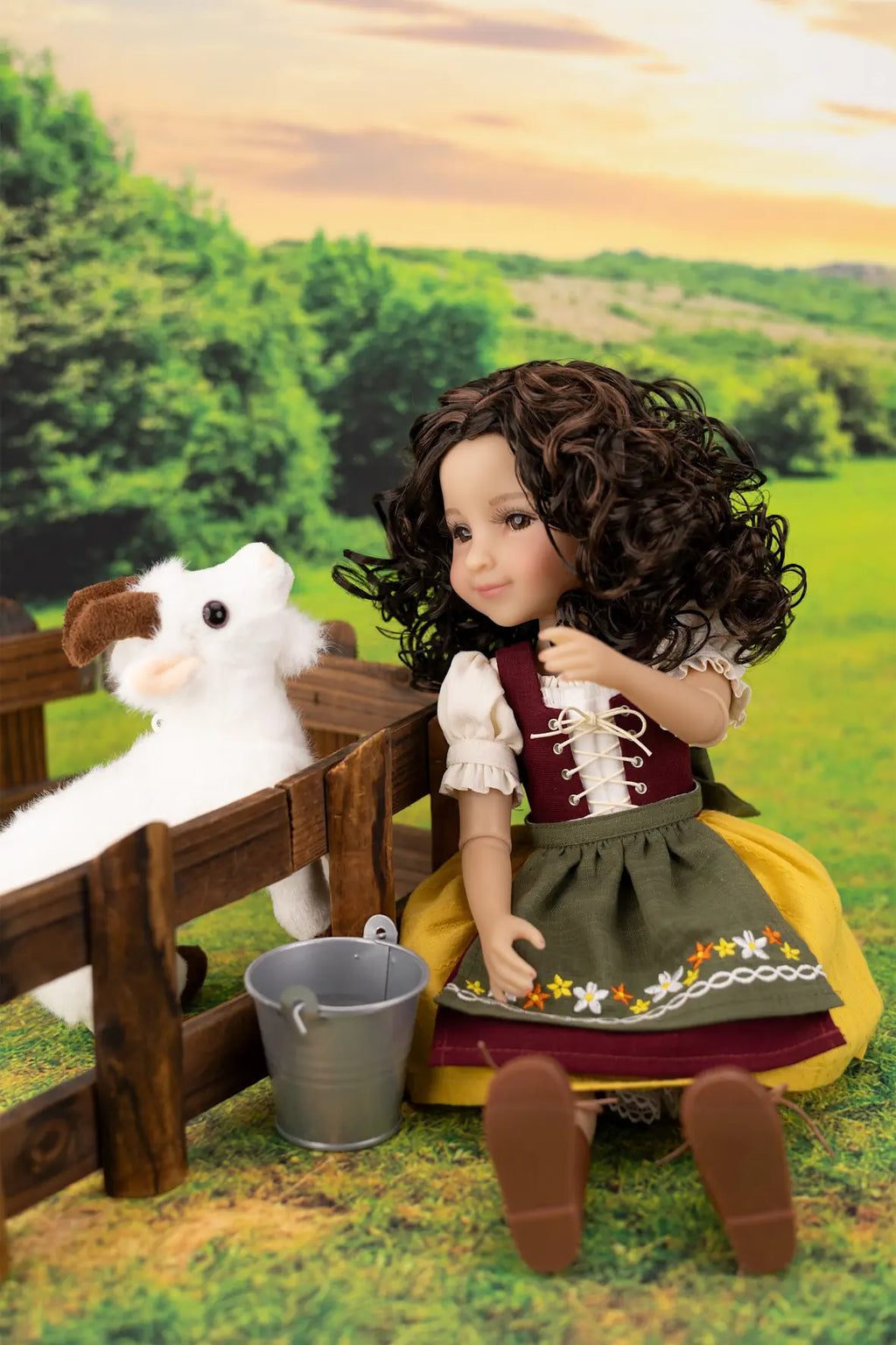 A doll with curly brown hair wearing a traditional German dirndl sits beside a small wooden fence in a staged outdoor farm scene, with a plush white cow and a metal bucket nearby.