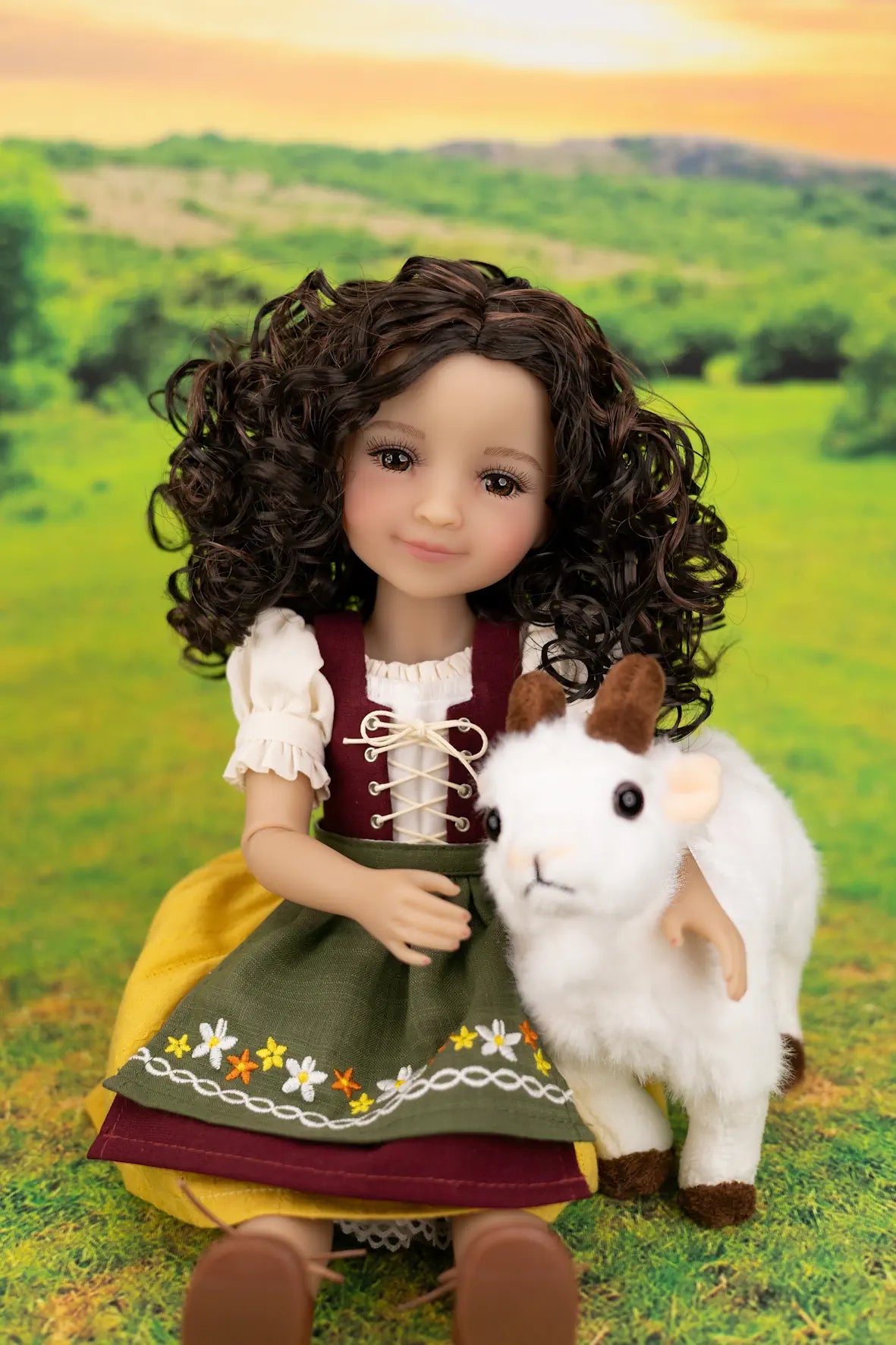 A doll with curly brown hair wearing a traditional-style dress, seated in a grassy field and holding a small white goat toy.