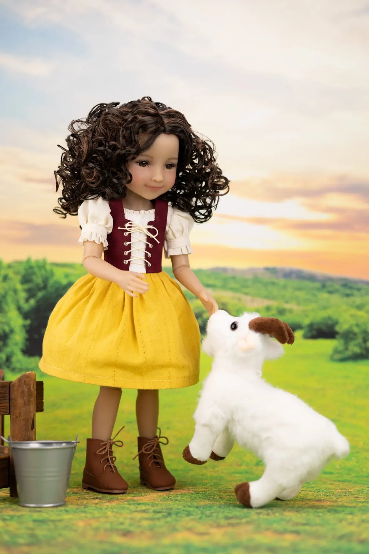A doll with curly brown hair wearing a yellow dress and brown boots, standing beside a small white plush lamb toy in a stylized outdoor farm scene with a sunset sky.
