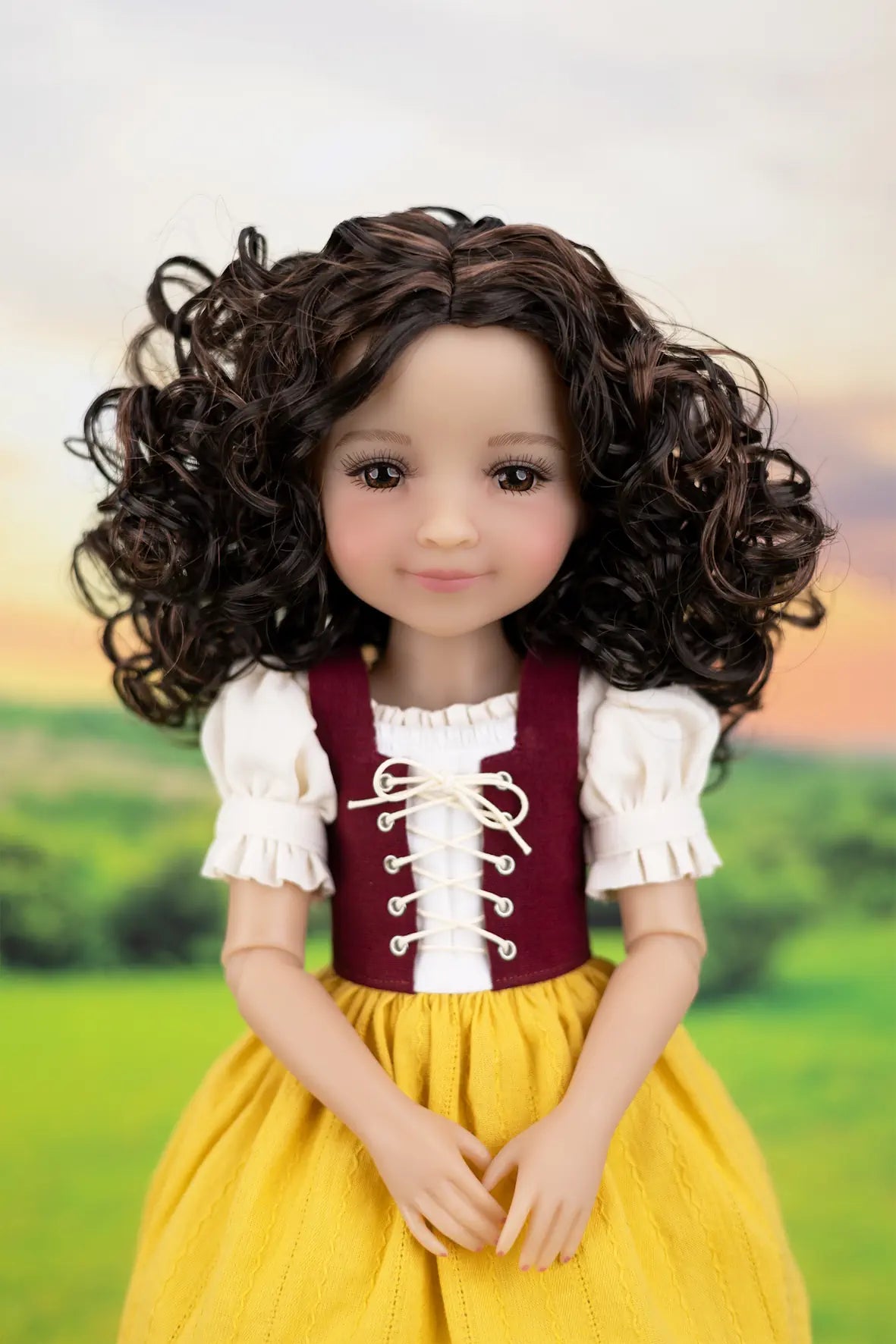 A doll with long, curly dark brown hair wearing a traditional-style dress with a maroon bodice, white ruffled sleeves, and a yellow skirt, posed against a blurred green landscape background.