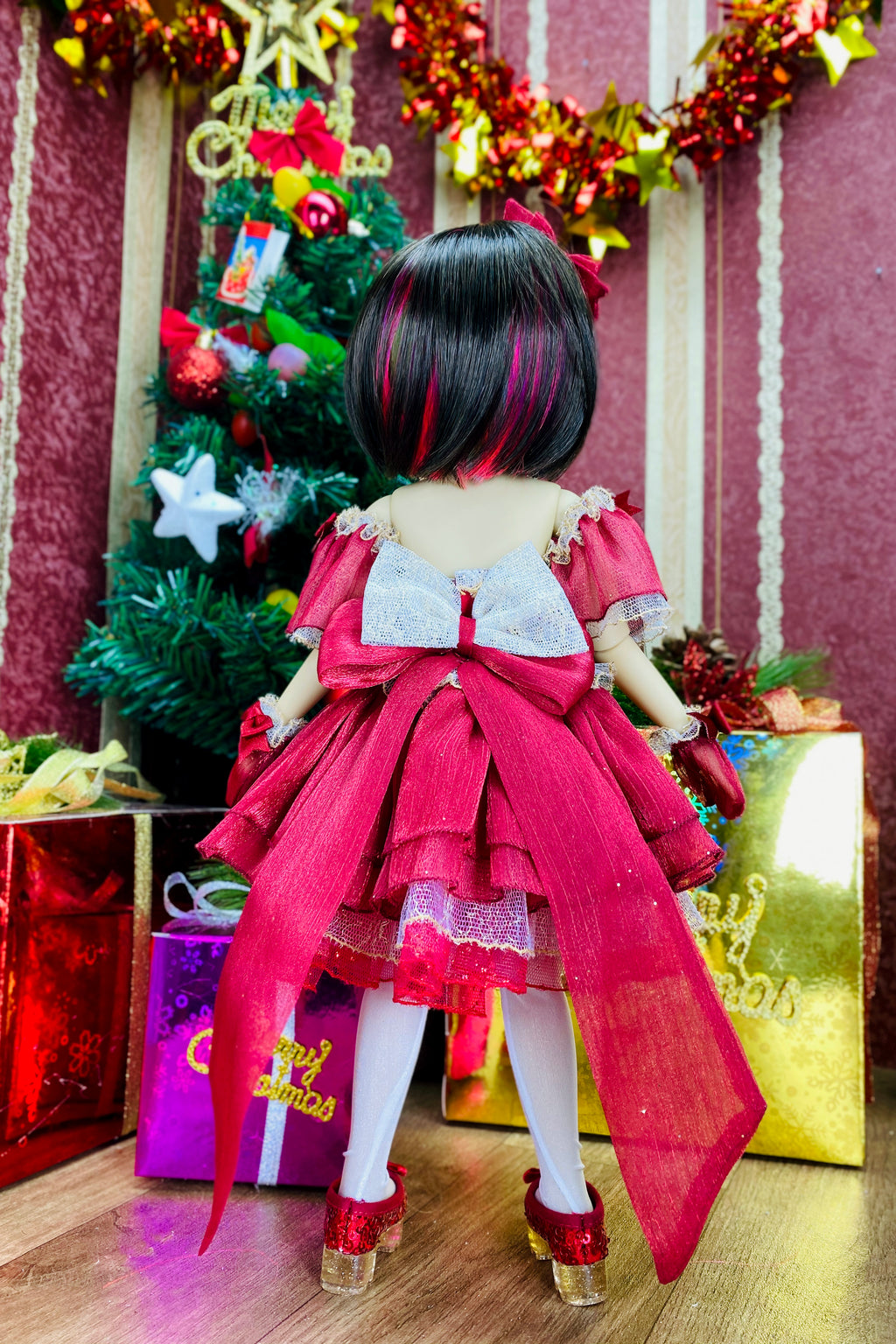 Doll in a red dress standing in front of a Christmas tree with decorations and presents.