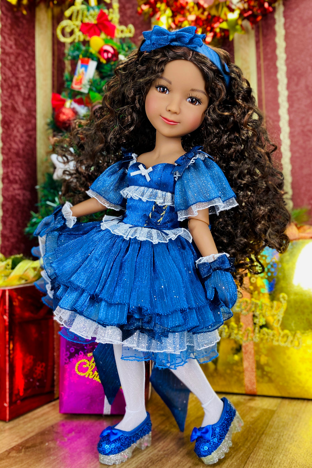 Doll in a blue dress with ruffles and a matching headband, standing in front of a decorated Christmas tree.