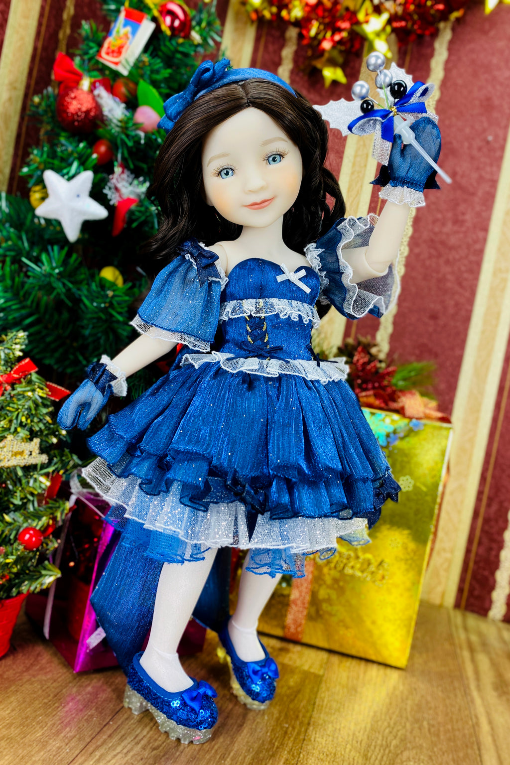 Doll in a blue dress with ruffles standing in front of a decorated Christmas tree.