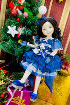 Doll in a blue dress standing in front of a decorated Christmas tree with presents.