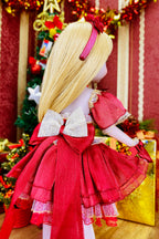 Doll in a red dress with a large bow, standing in front of a decorated Christmas tree.