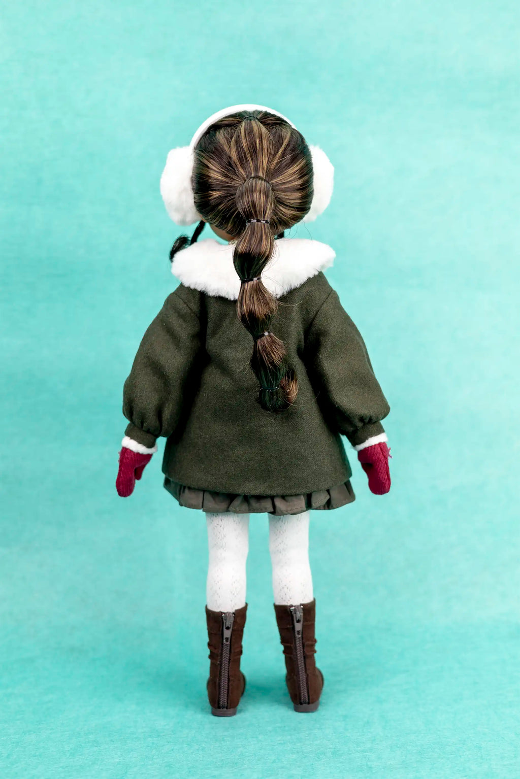 Doll wearing a green coat with white fur trim, brown boots, and white ear muffs on a light blue background.