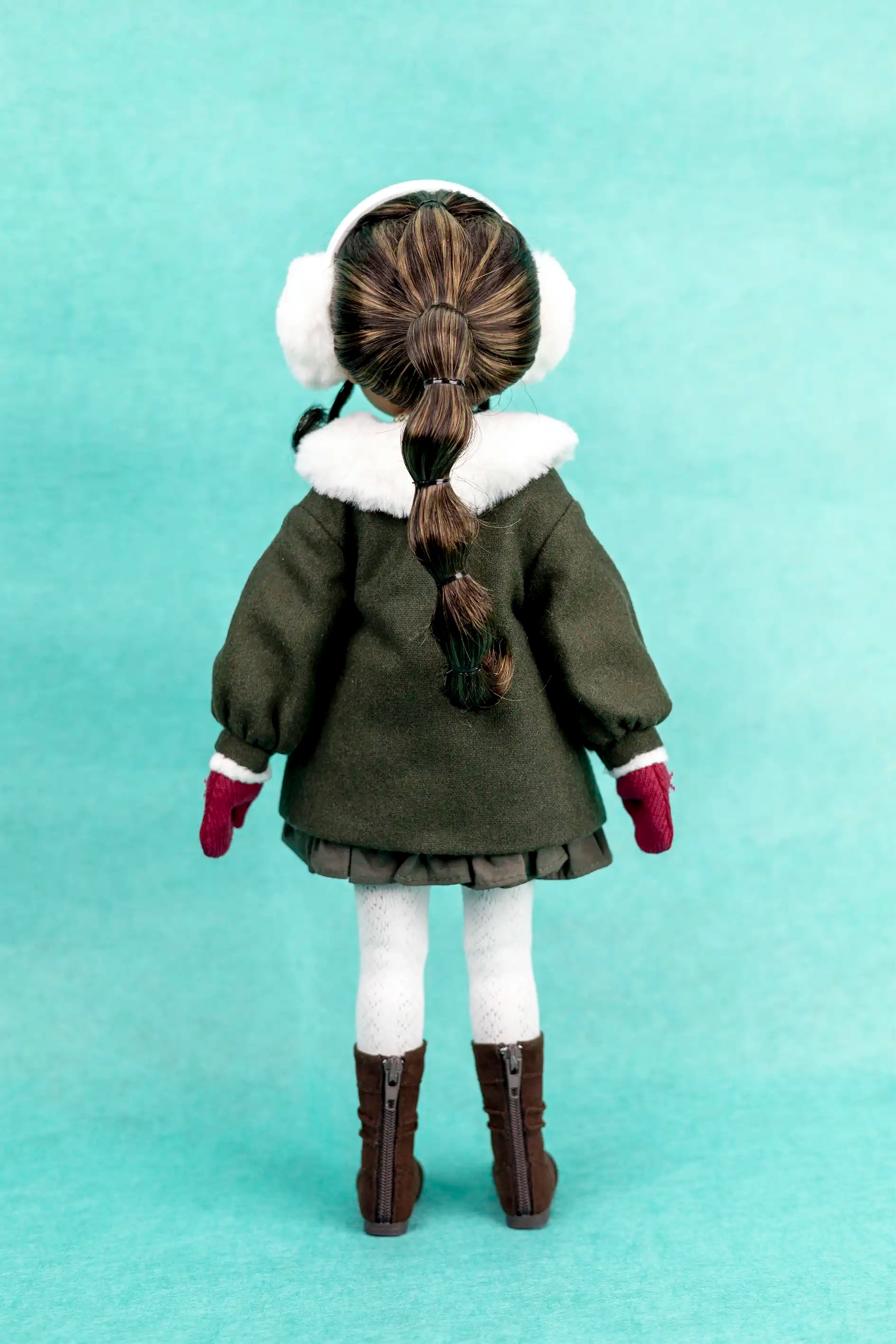 Doll wearing a green coat with white fur trim, brown boots, and white ear muffs on a light blue background.