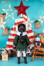 Doll in festive outfit standing in front of a decorated Christmas tree with lights and ornaments.
