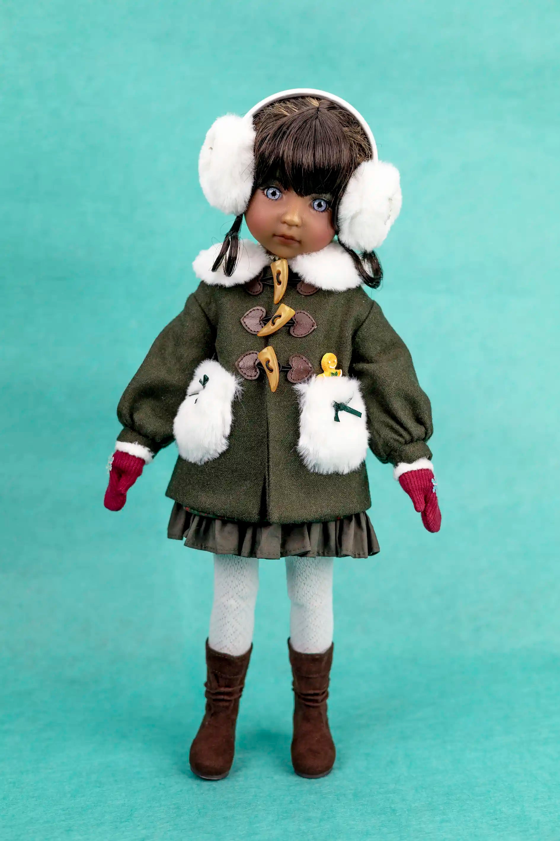 Doll wearing a green coat with decorative buttons, white gloves, brown boots, and white earmuffs on a teal background.