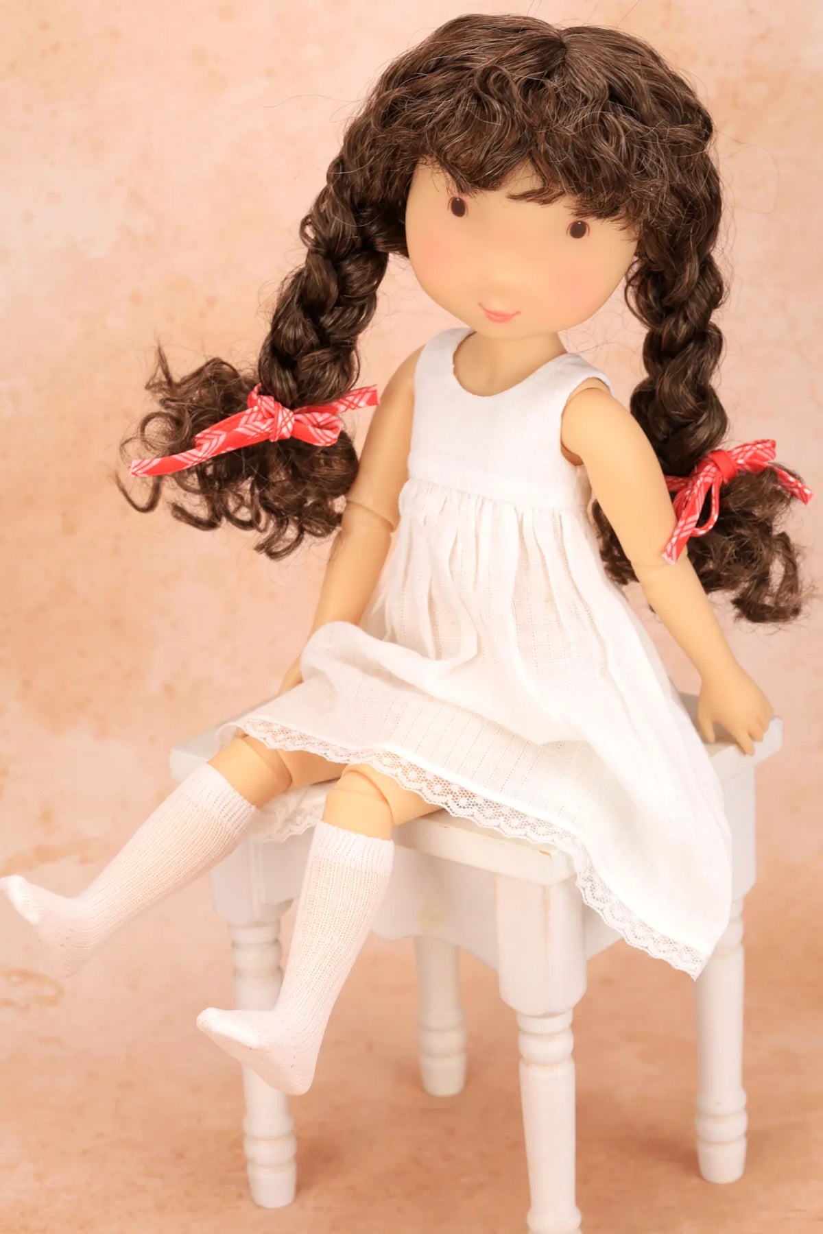 A Holly Hobbie doll with long curly brown hair tied with pink bows, wearing a white sleeveless dress and white socks, seated on a small white stool against a soft peach background.