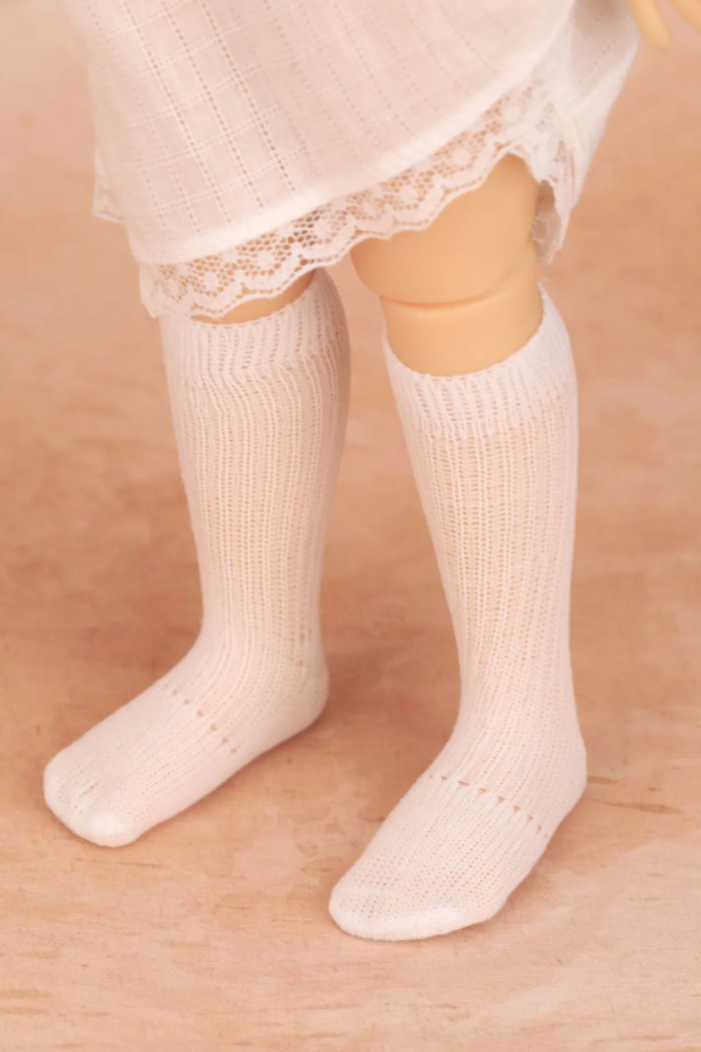 White ribbed knit knee-high socks worn on a doll's legs, with a white lace-trimmed skirt visible at the top, set against a warm beige background.