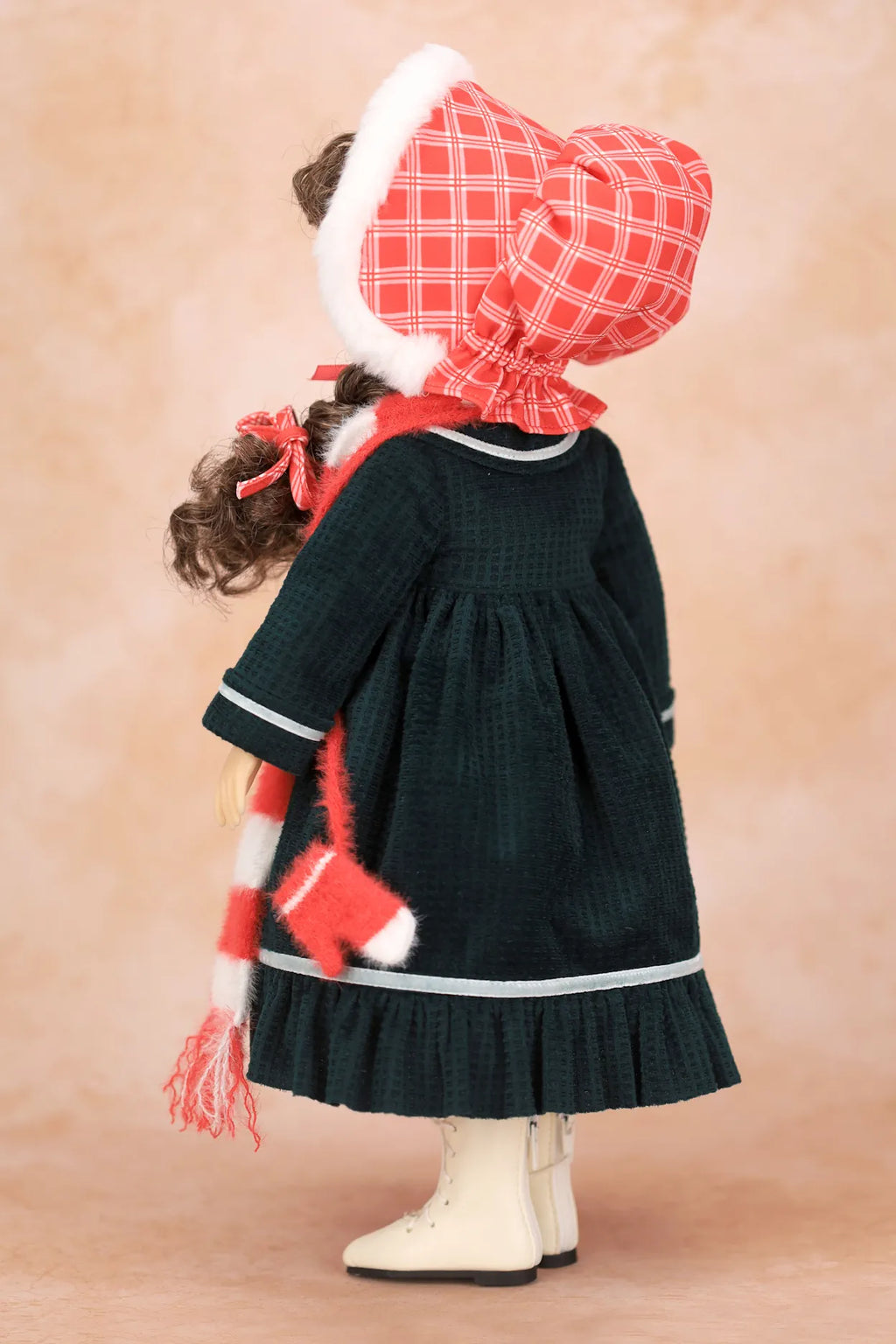 A Holly Hobbie doll viewed from the back wearing a dark green dress with white trim, a red and white plaid bonnet with white trim, a matching red and white striped scarf, and cream boots, set against a warm beige studio background.