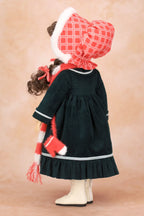 A Holly Hobbie doll viewed from the back wearing a dark green dress with white trim, a red and white plaid bonnet with white trim, a matching red and white striped scarf, and cream boots, set against a warm beige studio background.