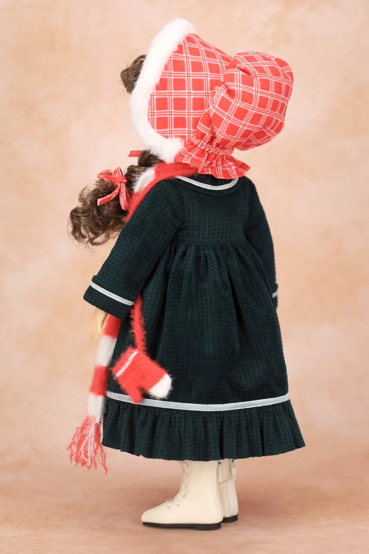 A Holly Hobbie doll viewed from the back wearing a dark green dress with white trim, a red and white plaid bonnet with white trim, a matching red and white striped scarf, and cream boots, set against a warm beige studio background.
