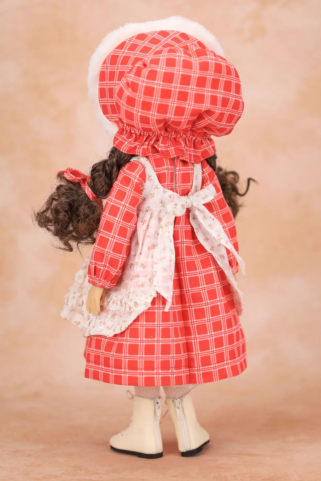 A Holly Hobbie doll viewed from the back wearing a red and white gingham dress with a white lace apron, matching bonnet, and white boots, set against a warm beige background.